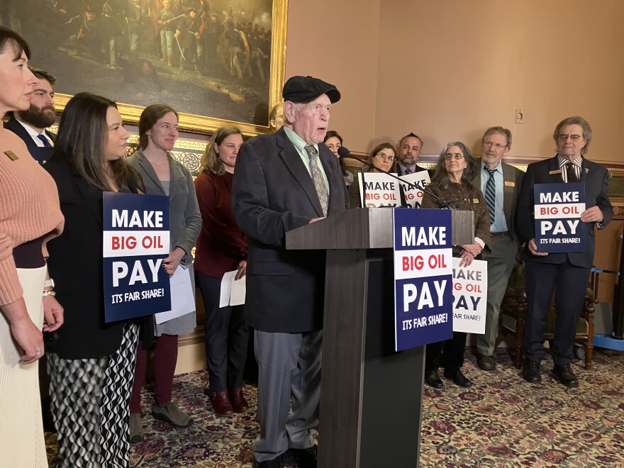 A man in a suit and a wool cap stands at the podium speaking, over a sign that reads 'Make Big Oil Pay It's Fair Share.'