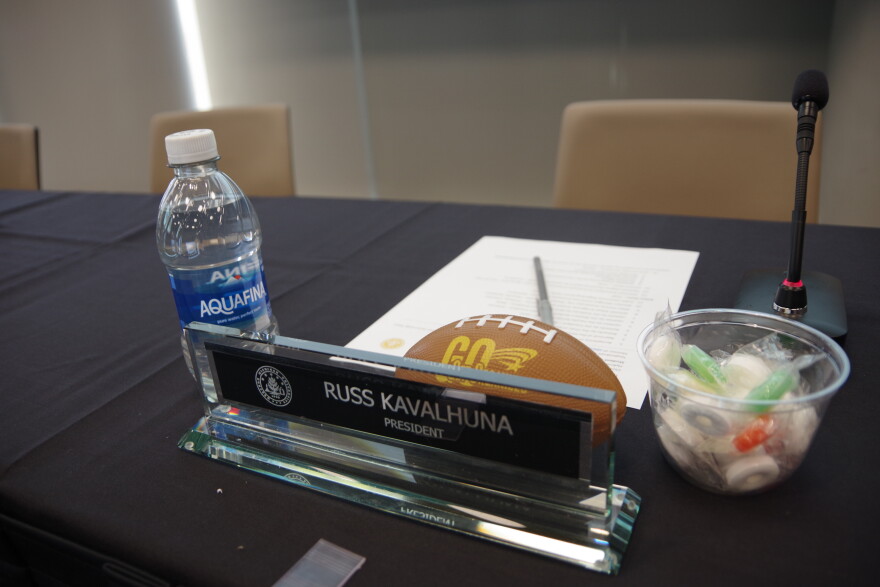 A nameplate that reads: "Russ Kavalhuna, President" sits on a black table. On either side of the nameplate are a bottle of water and a small plastic cup filled with Lifesaver candies. Behind the nameplate rests a small football-shaped stress ball that reads "get organized".