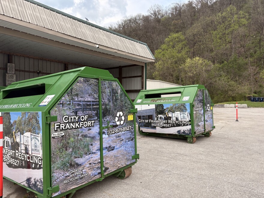 Collection bins at the City of Frankfort Recycling Center. The bins are about 5 feet tall with a footprint of about 4x7 feet. They're green, dumpster-shaped, with slits to receive recycling on both of the wider sides. Two of them are in the frame, sitting outside of an open covered space protecting additional bins from the weather.