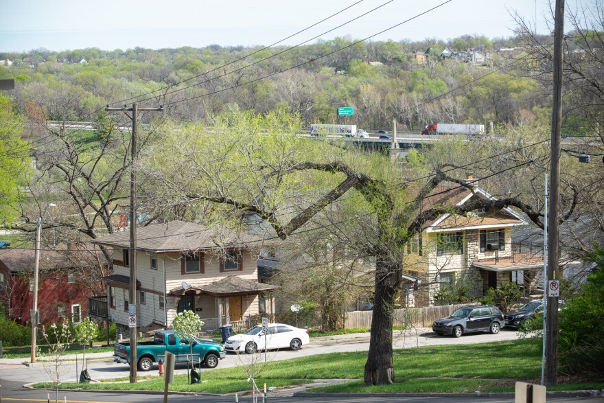 A pair of two-story houses sit in the foreground with cars parked in the street. In far background, a highway can be seen with traffic moving along it.