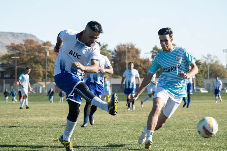 Action shot of two players from both teams facing off. To the left, an Afghan United player appears to have kicked the ball mid air while the Toofan player to the right runs in his direction. Blurry players from both teams are seen looking on the background.