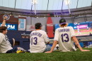 3 men in rays baseball jerseys sit on an astroturf field inside a stadium and watch a baseball game on a huge ballpark videoboard