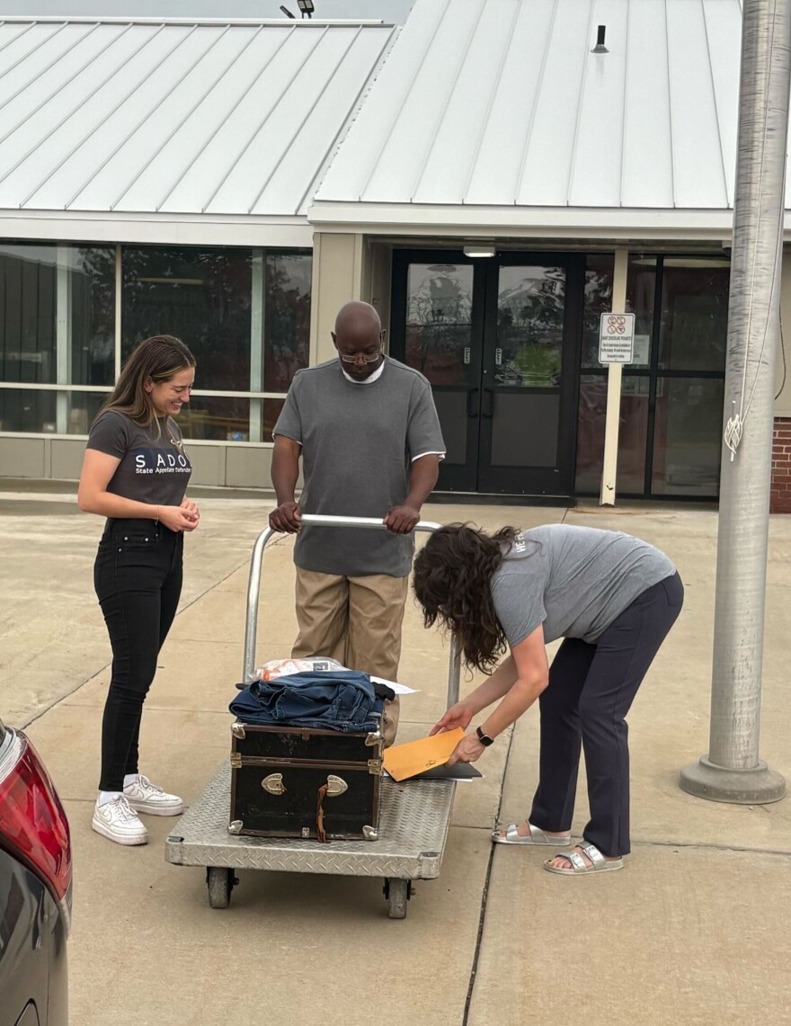 A man is standing outside a building, he's holding the handle of a metal cart that contains a few personal effects. Two women are standing on either side of the cart. One can be seen smiling, while the other is bent over to place what appear to be documents on the cart.