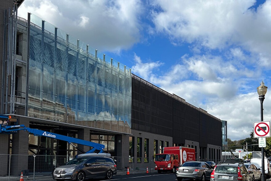 A large blue crane parked in front of an unfinished building with glass paneling and cars parked on opposite ends of the street.