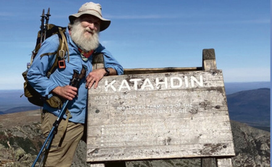 Man stands next to a sign stating "Katahdin" at the top of a vista. He is wearing hiking gear.