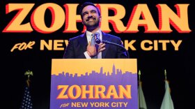Zohran Mamdani standing and smiling at a podium with a microphone, against a backdrop featuring the name "Zohran." The podium and background both display the text "Zohran for New York City." Several flags are visible in the background.