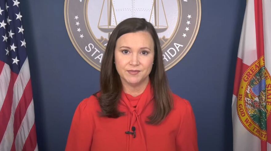 Florida Attorney General Ashley Moody stands to look at the viewer. Behind her to the left is the American flag and to the right, is the Florida flag. Directly behind her is a seal that reads "State Of Florida." Moody wears a jacket.