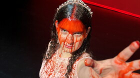 Young woman in pink dress wearing crown covered in fake blood looks up to the camera reaching her bloody hand up 