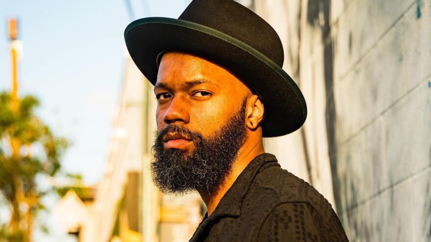  A man with a stylish hat and full beard looks at the camera while standing alongside the exterior of a cinder-block building.