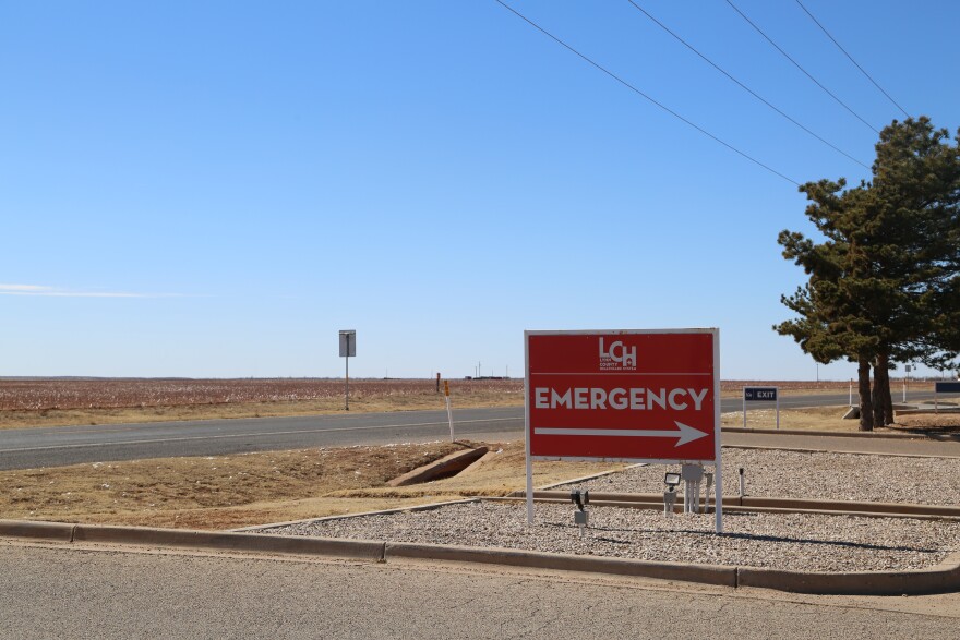 A photo of a red sign pointing to the emergency entrance to the Lynn County Healthcare System's hospital stands beside a long stretch of road and across from an empty field.