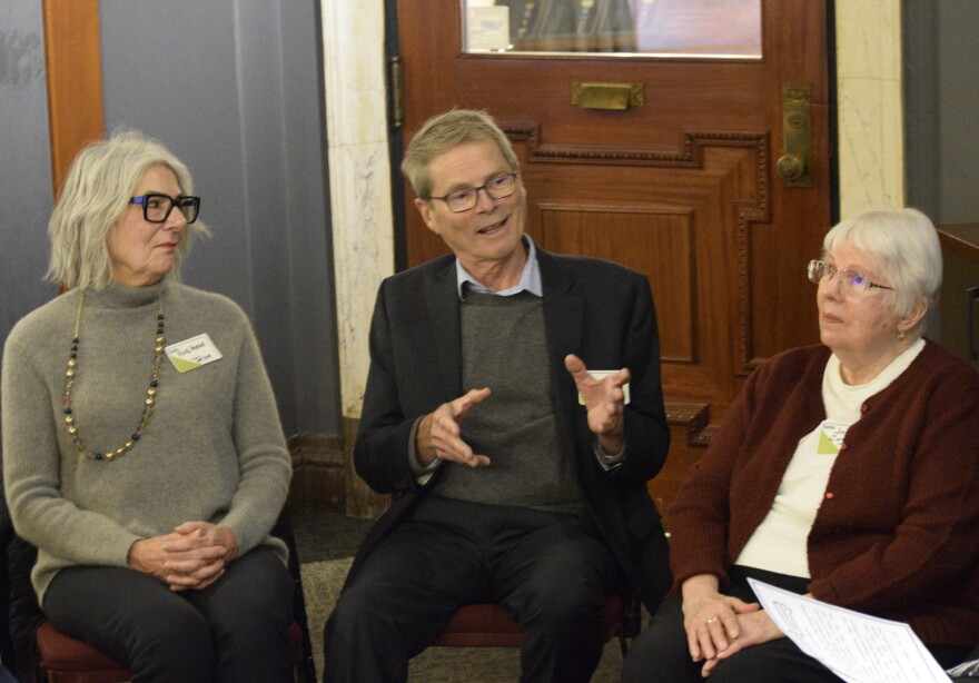 Three people sit on folding chairs, as part of a dialogue circle.  On the left is an older woman, with gray hair. In the middle, a man wearing a sweater and blazer, gestures while he talks. Another older woman with short white hair, and wearing a red cardigan, sits on the the other side listening.