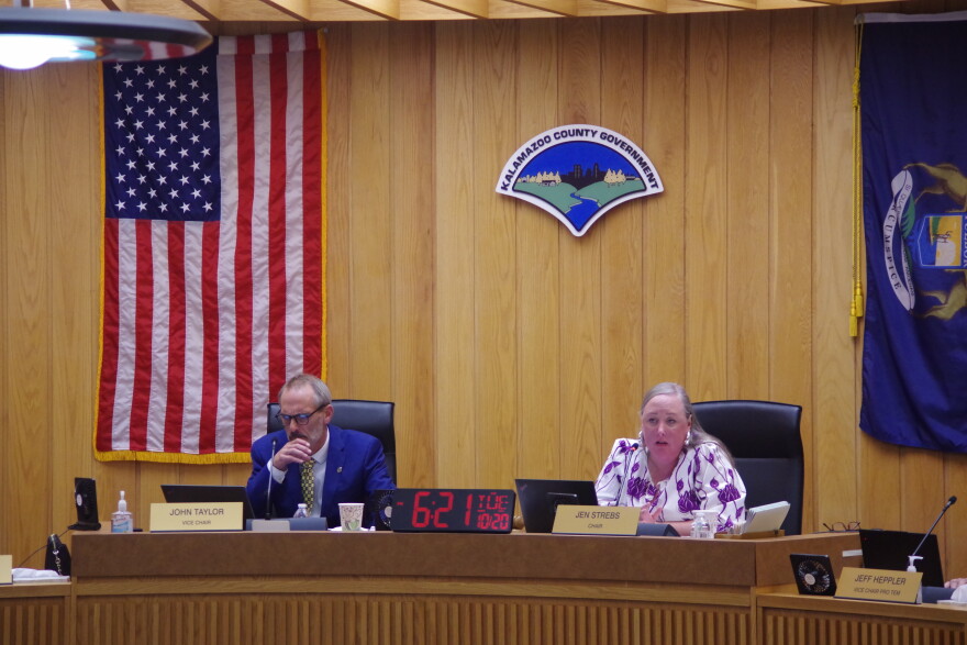 A man in a blue suit and yellow tie and a woman in a white and purple blouse sit on the left and right, respectively, of a long desk with mics and name plates to denote their positions as county commissioners. An American flag hangs on the wall on the left, while a Michigan flag hangs on the right. 