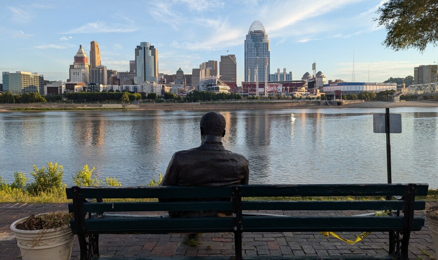 A bronze statue on a bench faces the Cincinnati skyline from Covington, Kentucky.