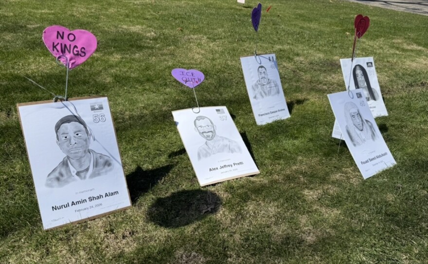 On a patch of grass, signs with drawings of faces along with names and dates of death are stuck in the ground. The posters depict high-profile cases of people who died after having contact with Immigration and Customs Enforcement -- one of the posters depicts Alex Pretti. The posters have construction paper hearts stuck to them with phrases like "no kings" and "ice out". 