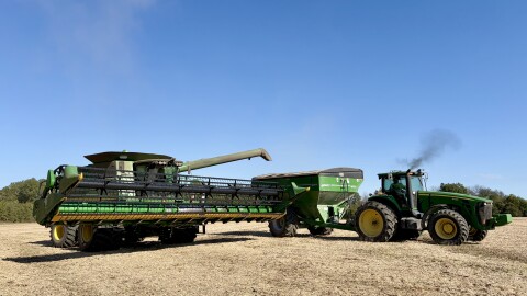 A green John Deere combine unloads soybeans into the wagon hitched to a green tractor. 