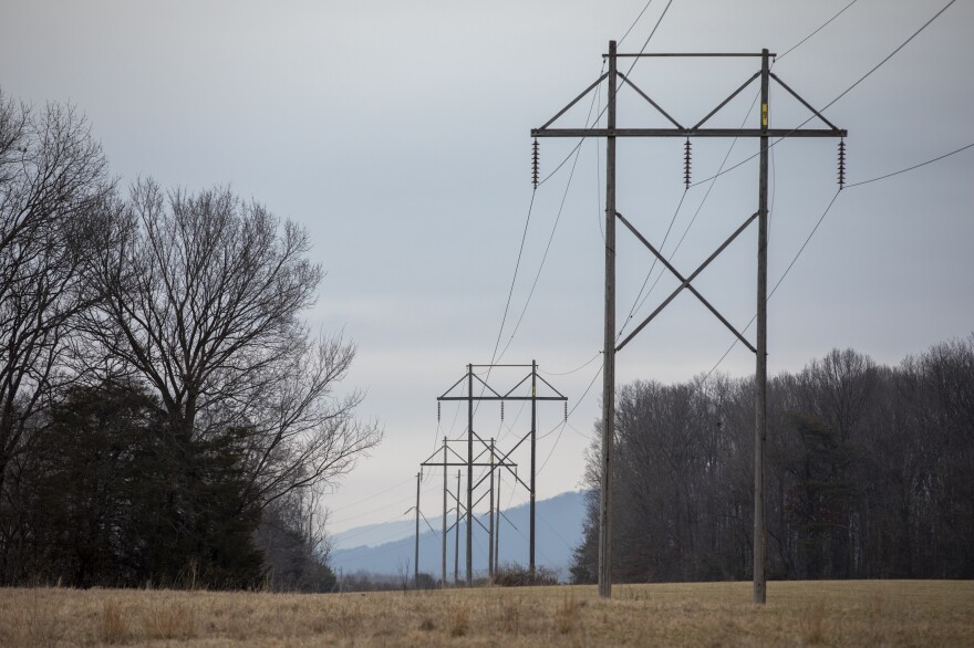 High voltage power lines run over a field with some trees to either side. 