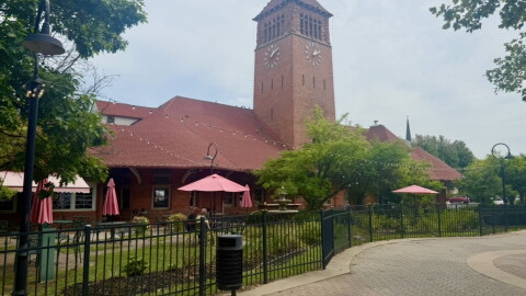 A large red brick building can be seen on a cloudy day. There is a large, fenced-in courtyard in front of he building with a grassy lawn. ON the front of the building is a large turret with a clock face on it. 