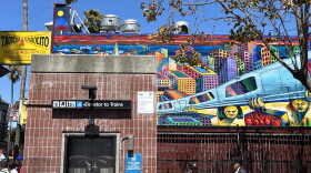 A mural on the wall of El Farolito on Mission Street, facing the northeast corner of the 24th Street BART plaza, by artists Michael J. Rios, Richard Montes, and Tony Machado. Depicts golden figures holding up a silver BART train against the colorful backdrop of the city. 
