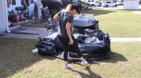 Dawn Gilliam, a white woman in her forties, next to a pile of trash bags on her front lawn. Gilliam is looking away from the camera at the pile, gesturing at the broken down table underneath it with her foot. 