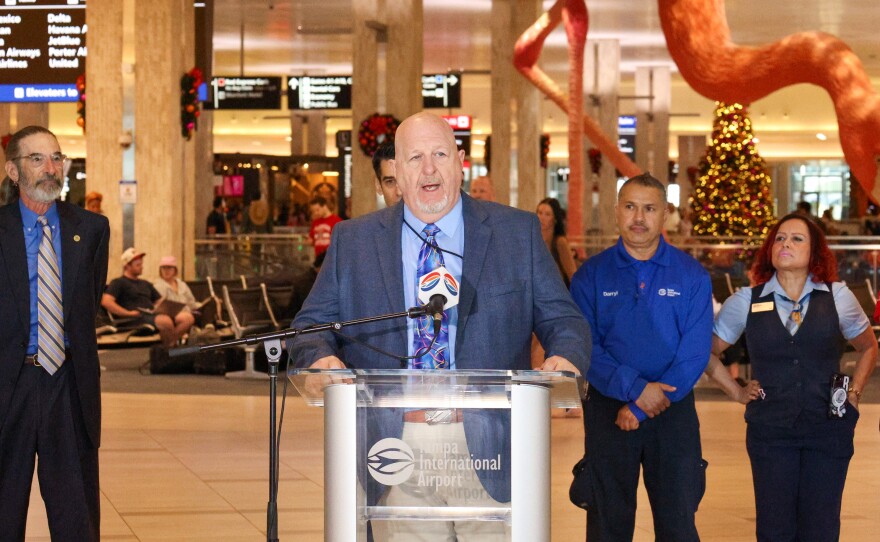 Man in a suit stands at a podium. Other officials, travelers, and a giant fiberglass and resin flamingo are behind him.