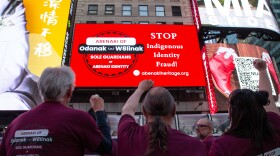 Members of Odanak and W8linak First Nations face a billboard they sponsored in Times Square in New York City during a United Nations Conference on Indigenous Identity Fraud. They wear red shirts and the billboard reads: "STOP Indigenous Identity Fraud! Abenaki of Odanak and W8linak, sole guardians of Abenaki Identity."