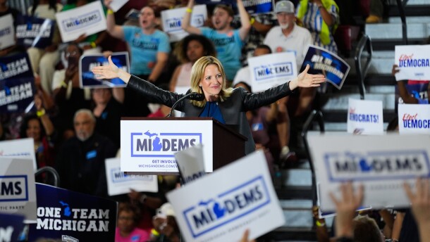A woman behind a stands with her arms extended out as a crowd of people hold campaign signs surround her.