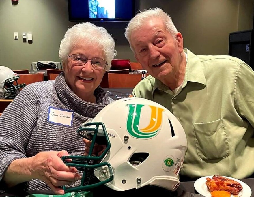Woman and man seated next to each other with a white football helmet between them that has a large green and gold "U" logo on the side.