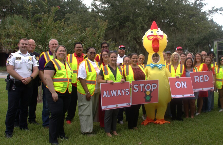 A group of people in neon traffic vests pose for a photo. They hold signs that read, Always Stop On Red. A police officer in uniform and a person wearing an inflatable chicken suit are also part of the group.