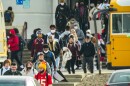 High school students, some masked and some not, walk out of a school building between two yellow, parked school buses.