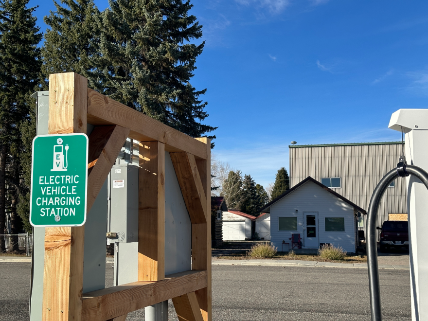A sign bolted to a wooden structure, reading "electric vehicle charging station." Part of the charging station is visible. Across the street are some small houses and a larger commercial building.