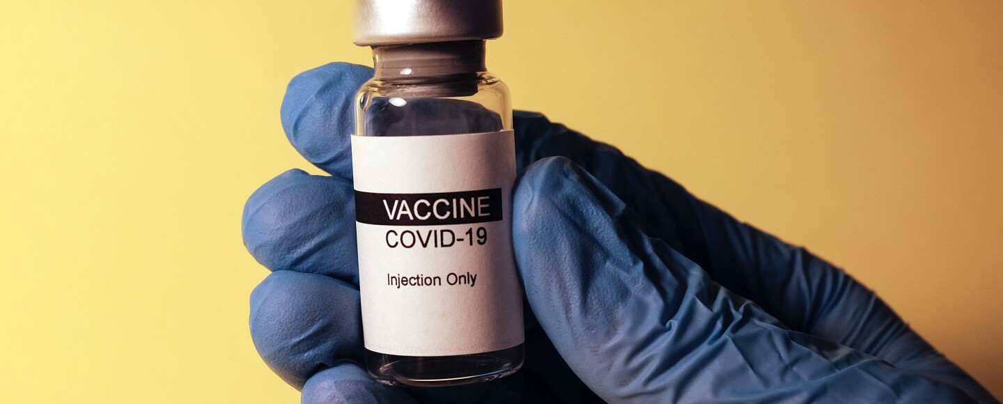 A hand wearing a medical glove holding a COVID-19 vaccine bottle.