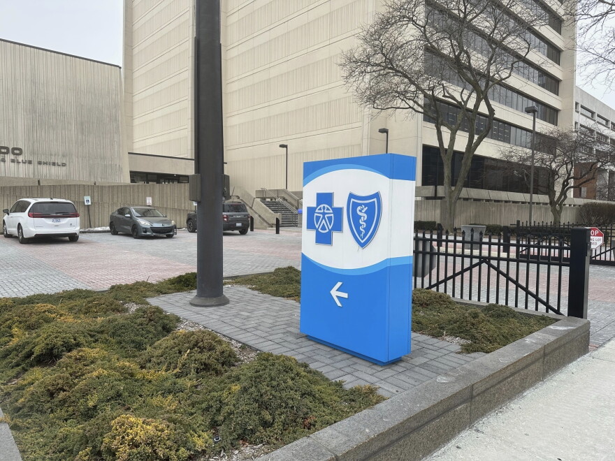 A blue and white sign with the Blue Cross Blue Shield logo stands in a grassy patch in front of a large grey building.