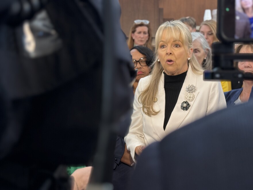 A woman appears to be mid-word as she looks at some cameras in the foreground. She has blonde hair and is wearing a black shirt under a white jacket. She is flanked by a number of people who are looking on seriously.