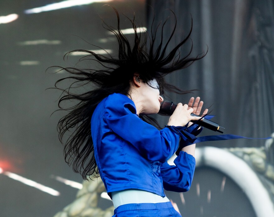A woman wearing a blue jacket sings into a microphone and dances so that her hair is flying over her head.