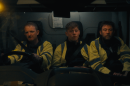 Three men in black shirts and reflective yellow jackets sit in the front seat of a utility vehicle. The one on the far left of the photo has his hands on the wheel, as the vehicle is from Ireland. All three men have serious facial expressions.