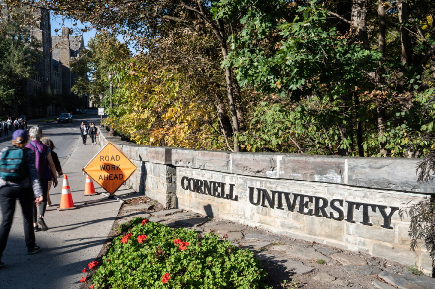 Students walk through campus on a sidewalk next to a long granite sign that says Cornell University 