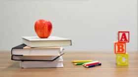 Four books stacked on a desk with an apple on top, next to some colored pencils and letter blocks