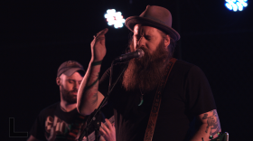 Two men illuminated on a stage. One holds a guitar in the foreground. The other signs into a microphone.