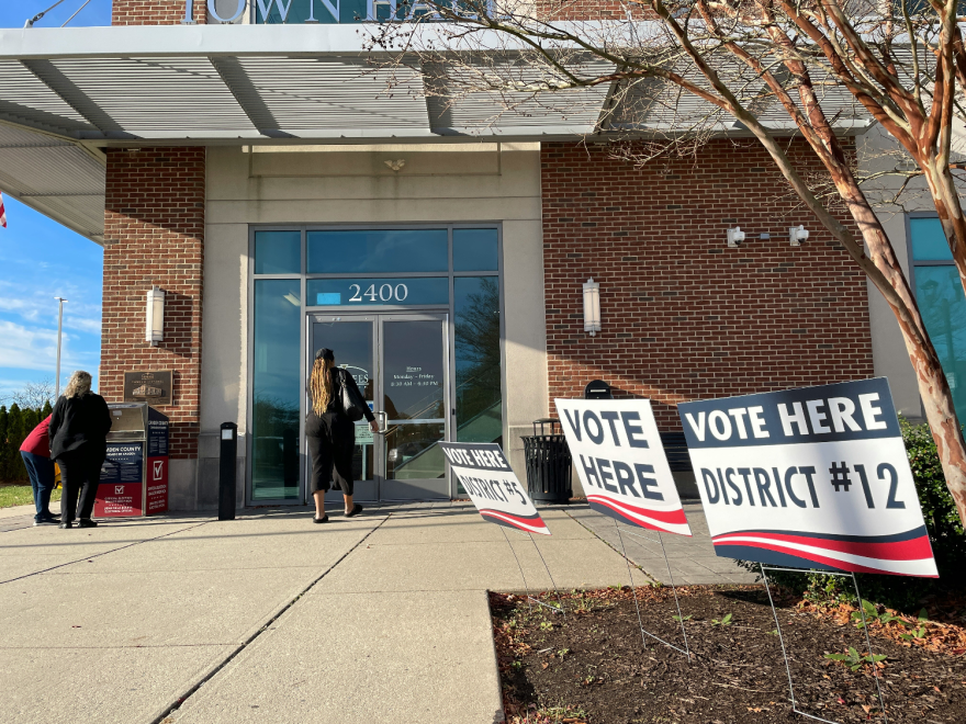 Photo of the outside of a  town hall. There are several signs saying "vote here" for different districts. One person is walking inside while two people are standing near a ballot drop box.