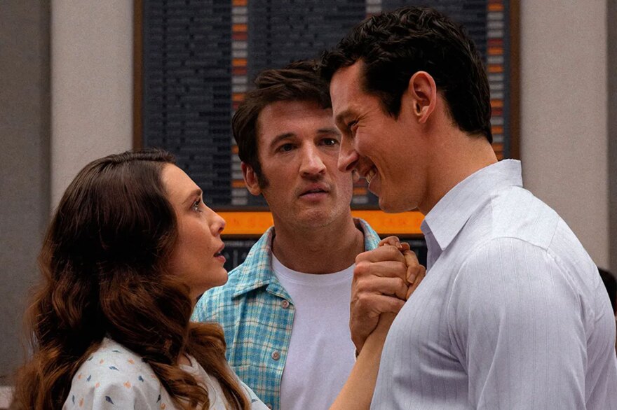 A still from the film Eternity. Elizabeth Olsen’s Joan and Callum Turner’s Luke stand close, hands clasped while Miles Teller’s Larry looks on. 