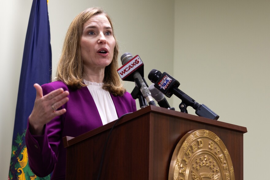 A woman in a purple blazer and white blouse speaks at a wooden podium with several microphones secured to it.