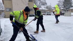 Three black men dressed in bright safety gear shovel snow in a driveway. The ground is covered with snow.