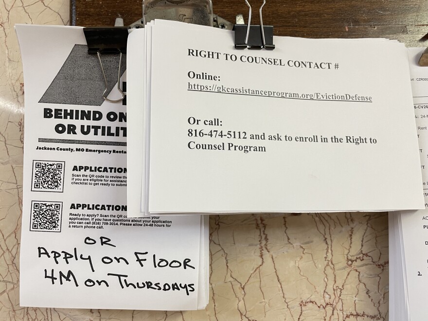 Stacked sheets of paper are hung next to each other on a wall. One of them reads "Right to Counsel Contact Number." The other says "Behind on rent or utilities?"