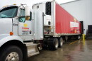 A large semi truck is parked at a warehouse. The trailer is completely red with a large Harvesters logo pasted in the middle of it's face.