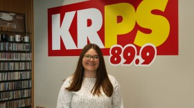 Riley Siegenthaler poses for a photo in the KRPS office. She's standing in front of a wall that has the KRPS 89.9 logo on it.