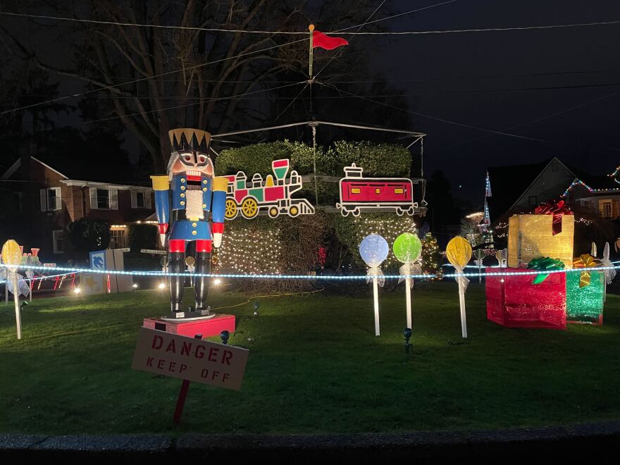 A larger than life-size nutcracker stands in front of a holly bush that has a cut out wooden train that circles around it. There are also three lollipops staked into the ground and beside those are three giftboxes stacked in a pyramid. This is all in the center of the neighborhood. 