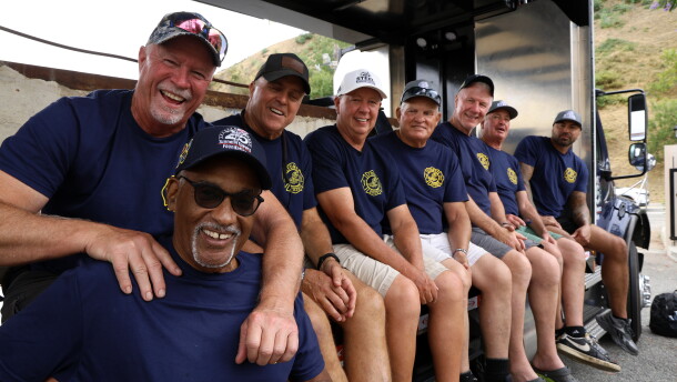 A row of men in blue T-shirts sit on a truck bed