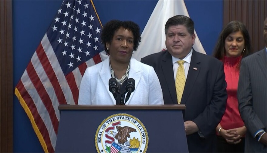A speaker stands at a podium with the Seal of the State of Illinois, flanked by two people. JB Pritzker addresses the crowd, highlighting reproductive rights. An American flag and another flag are displayed behind them.