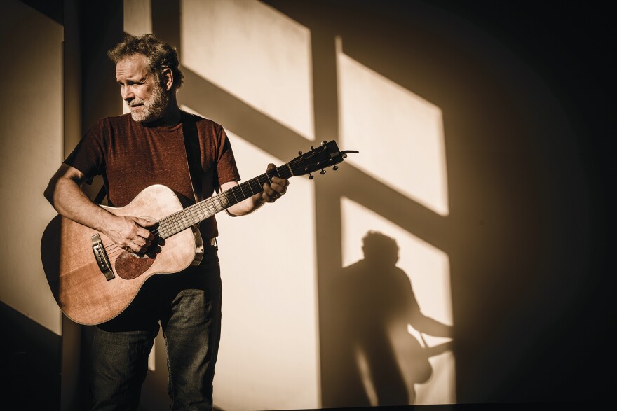 John Gorka stands illuminated by by sunlight from a window. He is holding a guitar and you can see his shadow against the wall behind him.