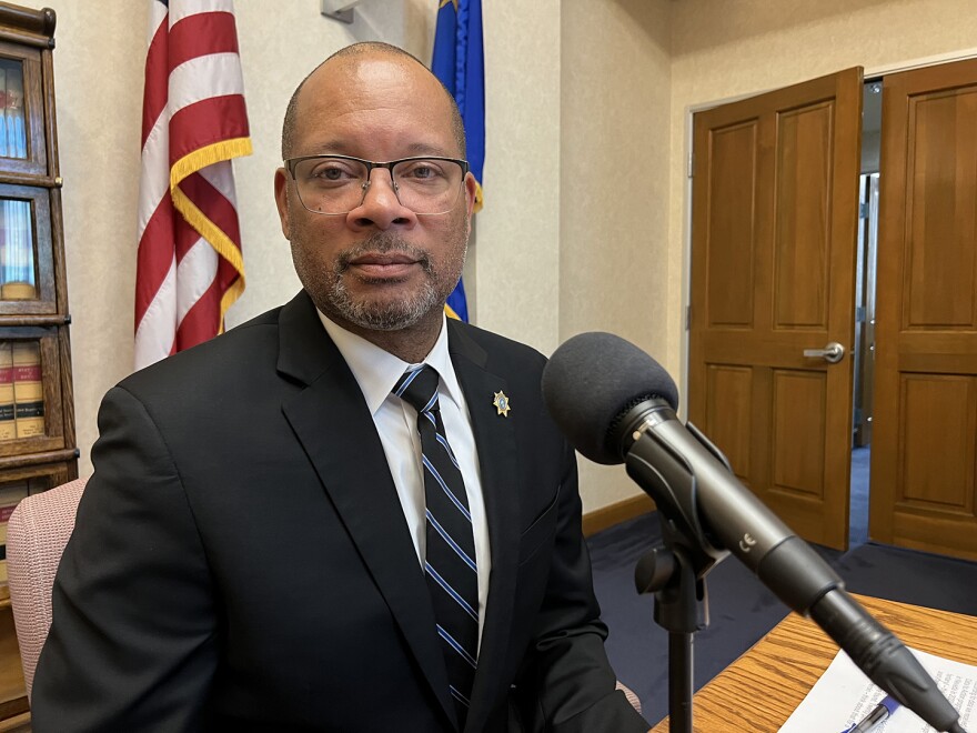 A man sitting at a table with a stern expression and a microphone pointed toward him. There is an American flag and a Nevada state flag on polls behind him.